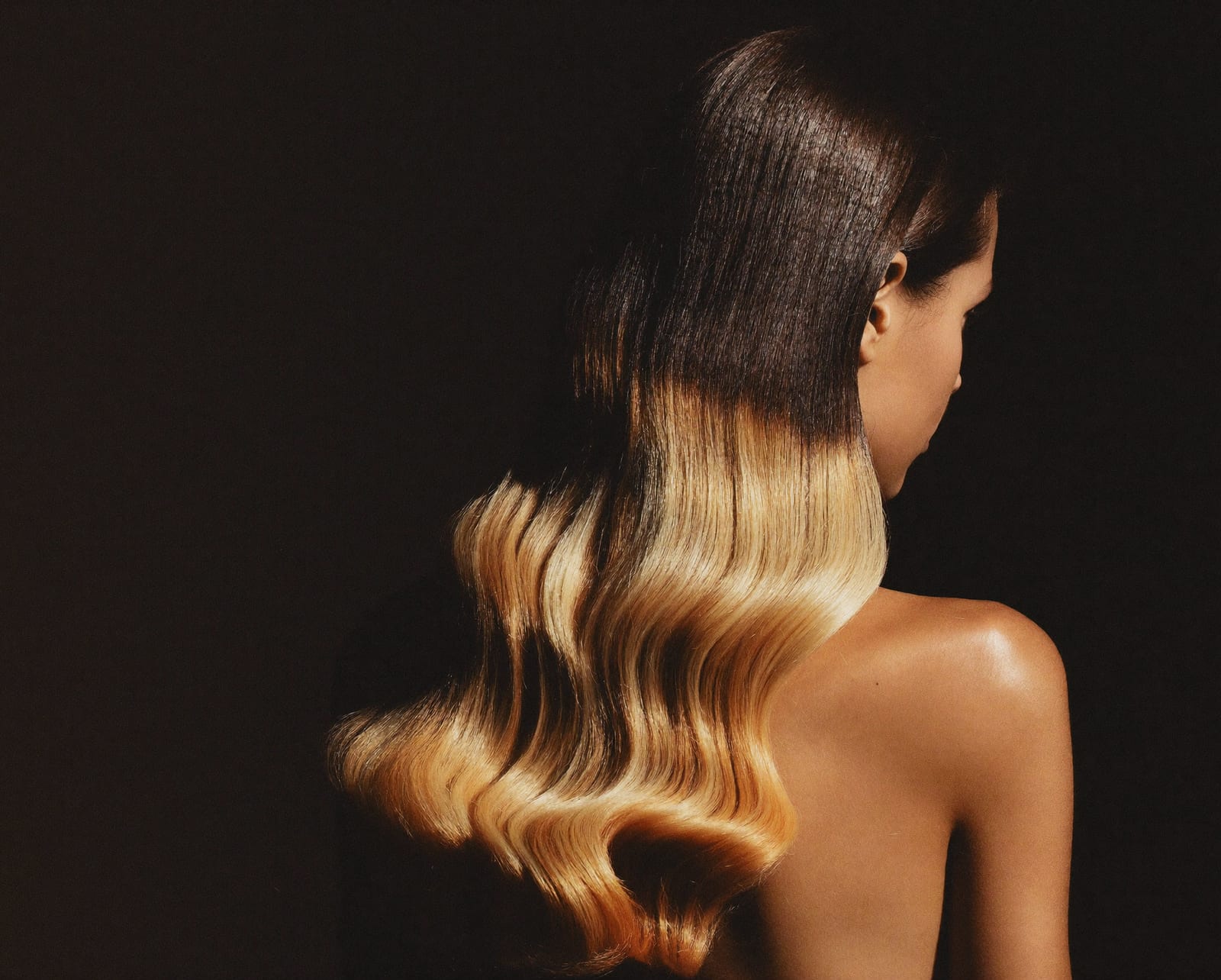 Long hair in a brunette-to-champagne colour melt, falling in polished waves, side-lit against a near-black background.